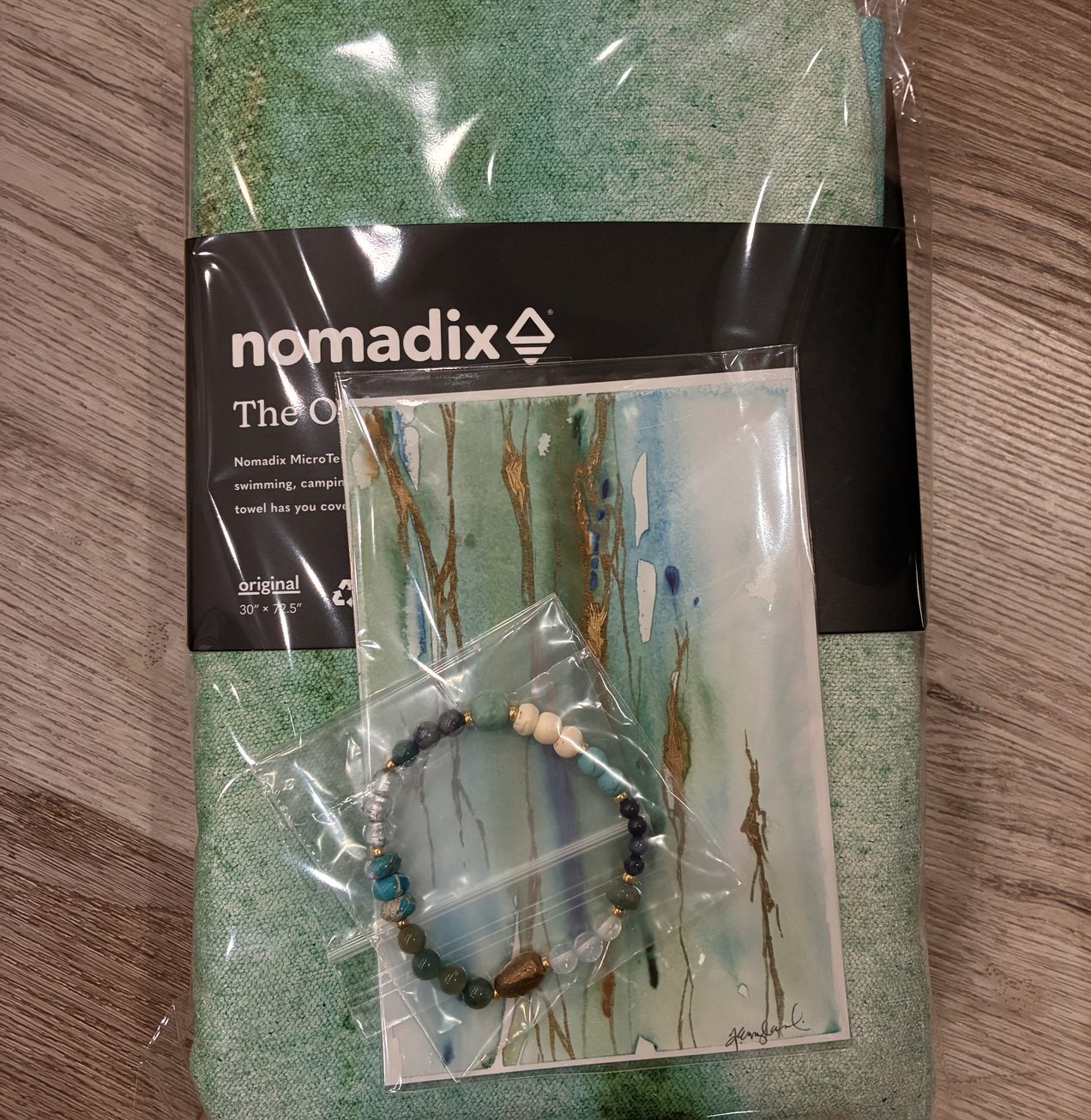 Nomadix Original Towel in its bag with the Illumination art card and a handmade bracelet